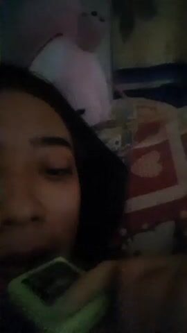 A woman is lying on her back in bed, partially obscured by a dark hood or head covering. She holds a small, lime green device with a glowing screen in front of her mouth. The device appears to be illuminated with green light. The background includes a patterned pillow with a Hello Kitty design, a partially visible pink stuffed animal, and a multicolored blanket.