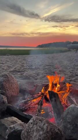 The video shows a campfire on a sandy beach at sunset. The sky is a mix of orange, pink, and yellow hues. A calm body of water is visible in the distance, with a treeline on the far shore. The campfire is made of logs and branches, with flames and glowing embers. Rocks are arranged around the fire pit. The sand is lightly textured, and there appears to be some grass or reeds in the background. The camera angle is low, close to the ground, focusing on the fire.