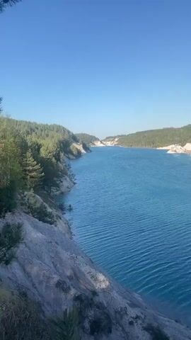 The video shows a body of water from an elevated perspective. The water is a vibrant blue, bordered by lush green forests on either side, which are dotted with some white, rocky outcroppings. The sky above is a clear, unblemished blue. Gentle ripples disturb the surface of the water, adding texture to the scene. In the foreground, a rocky cliff edge is visible, scattered with small trees and greenery. The overall impression is one of natural beauty and tranquility.