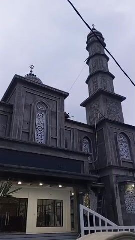 A low-angle shot shows the exterior of a mosque under construction. The building features a tall minaret, domes, and intricate window designs. The sky is overcast. Some construction materials and scaffolding are visible, along with some greenery. A few people are present, including one person on a ladder near the top of the minaret.