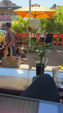 The video shows an outdoor cafe or restaurant area. In the foreground, a person's legs and lap are visible, suggesting they are seated at a table. In the background, a woman stands holding a baby. Several children's toys, including dolls and a toy car, are scattered on the ground near a wicker basket. A man walks by in the middle ground, pushing a cart. Other people are seated at tables under an orange umbrella and in the background. Lush green plants and trees are visible throughout the scene, and the overall environment appears to be a sunny day.