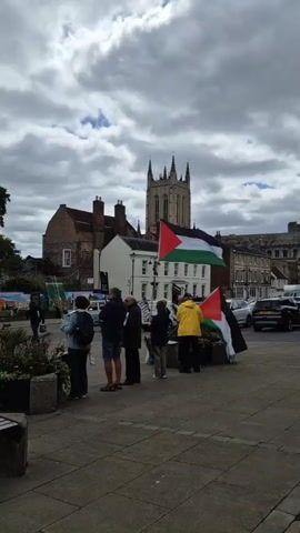 A group of people stand in a town square, some holding Palestinian flags. In the background, a tall, ornate church tower is visible, along with a stone building and other structures. A silver van drives by, and a person walks a dog on a leash across the square. The sky is cloudy.