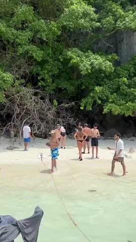 A group of people are gathered on a sandy beach with clear water. They are dressed in swim attire and casual clothing. Some are standing, while others appear to be walking or interacting. Behind them, there is lush green vegetation and rocky cliffs. The sun is shining, casting light on the scene. One person is holding what appears to be a small camera or phone on a stick.