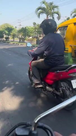 The video shows a street scene from the perspective of a motorcycle rider. In the foreground, the handlebars and part of the rider's hand are visible. Ahead, a red motorcycle with a rider wearing a black helmet is moving away from the camera. To the right, a yellow and green van is traveling in the same direction. The van has its side door open. Trees and utility poles are visible in the background under a clear sky. The road surface appears to be asphalt.