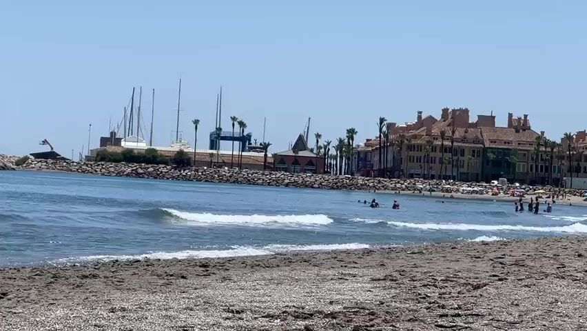 This is a wide shot of a beach scene. The foreground is a sandy beach with small pebbles. In the middle ground, the ocean has gentle waves rolling onto the shore. Several people are visible swimming and wading in the water, some closer to the shore and others further out.

The background features a harbor area with a breakwater made of large stones. Several sailboats with tall masts are docked in the harbor. Beyond the harbor, a row of multi-story buildings with terracotta roofs and colorful facades line the waterfront, suggesting a coastal town or resort. Palm trees are scattered among the buildings and along the shoreline.

The sky is clear and blue, indicating a sunny day. The lighting suggests it is daytime, likely late morning or early afternoon. The overall mood is relaxed and leisurely, typical of a beach vacation. There is no visible text in the media file. The scene appears to be in San Roque, Spain, as per the provided location context.