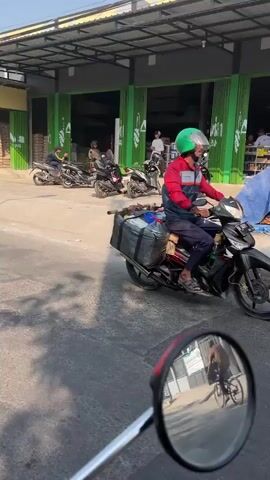 The video shows a street scene with several motorcycles. In the foreground, the reflection of a cyclist is visible in a motorcycle's rearview mirror. A person wearing a red and blue jacket and a green helmet is riding a motorcycle with a large package strapped to the back. Other motorcycles are parked in the background near a building with green doors. The environment appears to be outdoors during the daytime, with sunlight casting shadows.