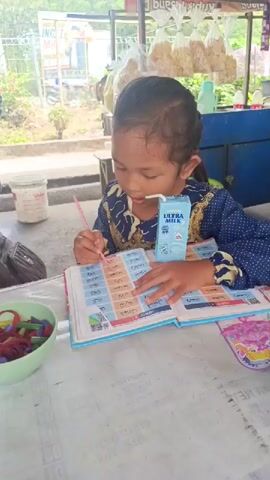 A young girl with dark hair tied in a ponytail is sitting at a table outside and is reading a book. She is wearing a blue and gold patterned dress. She is holding a pink pen and pointing at the book, which is open to a page with grids of text and images. A carton of milk with a straw is propped up on the book. To the left of the girl, there is a light green bowl filled with colorful hair ties. In the background, there are some food items hanging in bags and a white bucket. The environment appears to be an outdoor seating area, possibly at a roadside stall.