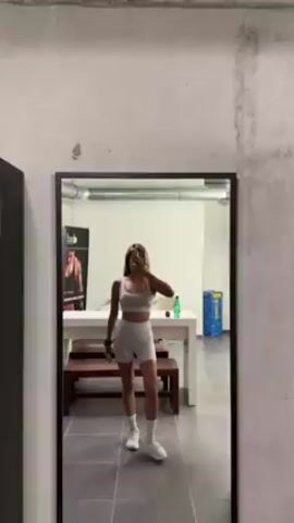 A slender person with dark hair and tan skin is taking a mirror selfie in what appears to be a gym or workout room. The person is wearing a matching gray sports bra and shorts set, with white sneakers and socks. The person is holding a phone in their left hand, partially covering their face.  Behind the person is a white table with a bench underneath, a water cooler, and a banner with a muscular person on it. The room has tile flooring and exposed ductwork in the ceiling.