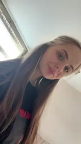 A young woman is looking at the camera from slightly above. Her hair is brown with blonde highlights and she is wearing a black shirt with red lettering. Music plays in the background. The scene is indoors with a window and curtains visible in the background.