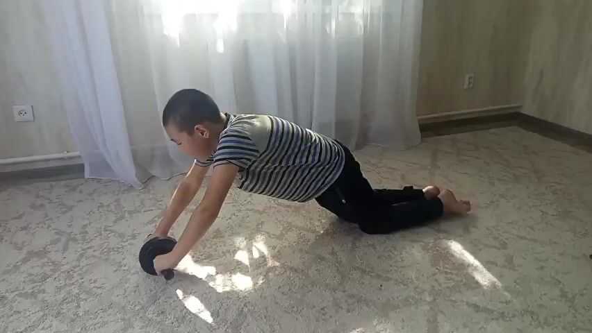 A boy is doing an ab roller exercise indoors. He is on his knees with the ab roller in front of him. He rolls the ab roller forward, extending his body into a plank position. He then rolls the ab roller back to his starting position. The room has carpeted floors and white curtains.