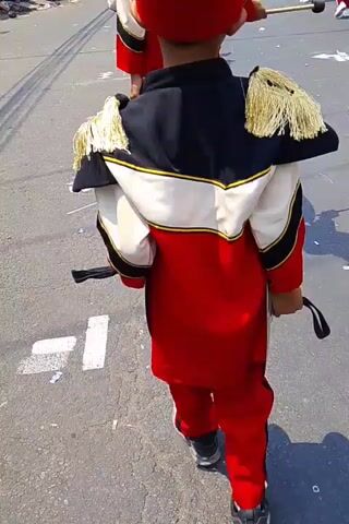 A first-person perspective video captures a child in a marching band uniform, complete with a tall red hat adorned with a fluffy pink feather, walking down a paved street. The child's uniform is red and white with black accents and gold fringed epaulets. They are holding a drumstick in their right hand. In the background, other band members in similar uniforms are visible, some also holding drumsticks. Spectators are lined up on the left side of the street, watching the parade. The buildings on the right side of the street appear to be shops. The sun is shining, creating shadows on the road.