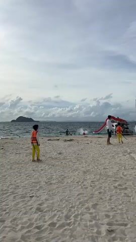 The video shows a beach with white sand. In the foreground, a child wearing a yellow shirt and pants, and an orange life vest walks towards the right. Further back, an adult male wearing a white shirt and dark shorts stands with a person in a yellow shirt and pants, also wearing an orange life vest. Behind them, a red inflatable slide is attached to a white structure, possibly a boat or a platform. The ocean is visible with waves and a distant island. The sky is cloudy. A bird is flying in the sky.