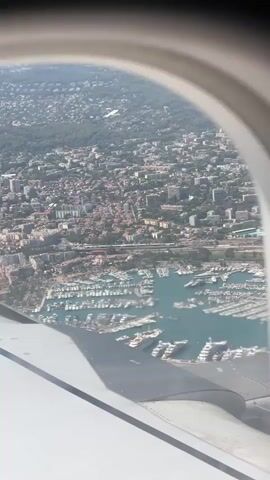 This is an aerial view from an airplane window of a coastal city with a large marina filled with boats. The city has many buildings and is built on a hillside. The wing of the airplane is visible in the foreground.