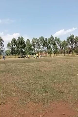 This media file shows a soccer game taking place on a grassy field. Several people, likely players, are scattered across the field, some in yellow shirts and others in blue and white. There is a soccer goal visible on the right side of the frame. The setting appears to be outdoors, with a line of tall trees forming a backdrop. The sky is blue with a few white clouds, suggesting it is daytime, likely late morning or early afternoon, with fair weather. The field itself is covered in dry, patchy grass, indicating it might be a rural or less maintained area. The overall scene depicts a casual game of soccer in an open, natural environment. There is no visible text in the media file. The location context provided is Lumakanda ward, Kenya.