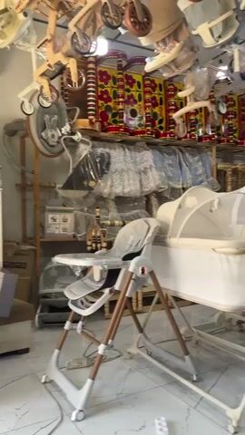 The video shows an indoor scene, likely a retail store specializing in baby products. In the foreground, a white baby bassinet with a mosquito net is prominently displayed on a stand. To the left, a grey high chair with wooden legs is visible. The background is filled with various baby items, including strollers hanging from the ceiling and stacked on shelves. Decorative textiles with colorful patterns are also visible on shelves. The lighting suggests it is daytime, and the floor appears to be tiled. There are no people or animals visible in the frame.