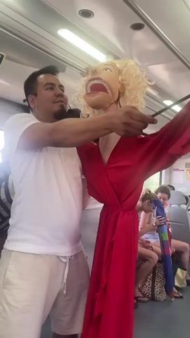 A man in a white t-shirt and light-colored shorts holds a large, blonde-haired puppet resembling Marilyn Monroe. The puppet is wearing a red dress with wide sleeves and has an exaggerated facial expression with its mouth open. The man uses a stick to manipulate the puppet's head and mouth. They are inside what appears to be a train car, with other passengers visible in the background. One woman is holding a colorful umbrella. The lighting suggests it is daytime.