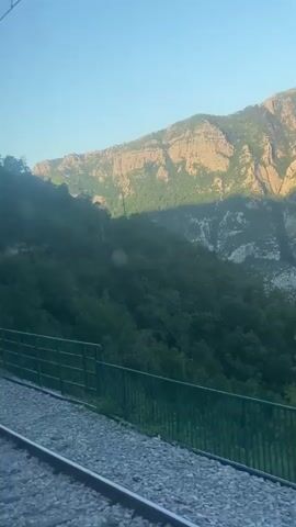 A video shows a train traveling along a track. The train is moving through a mountainous area with lush green trees covering the slopes. The sun is setting, casting a warm glow on the rocky peaks. In the foreground, the train tracks are visible, with a metal fence running alongside them. The camera is inside the train, looking out the window. The train is passing by a tunnel entrance in the mountainside. There are no people visible in the video. The overall mood is serene and scenic.