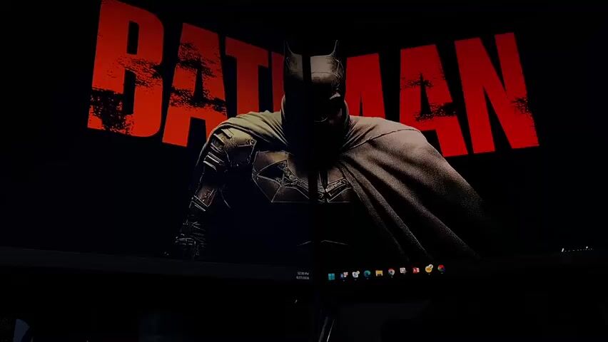 This media file is a still image featuring a computer monitor displaying a dark, moody wallpaper. The wallpaper shows a silhouette of Batman from the chest up, with his cape flowing around him. Above Batman, the word "BATMAN" is emblazoned in large, distressed red lettering.

The monitor itself is dark, suggesting the room it's in is dimly lit, possibly at night or in a controlled environment. The bottom of the screen shows a taskbar with several icons, including what appears to be a calendar indicating the date 4/17/2025 at 12:30 PM, and various application shortcuts. The computer is set up on a desk, and the reflection of the monitor is visible on what appears to be another monitor or a dark surface below it.

The overall mood conveyed by the wallpaper is one of intensity and darkness, fitting for the Batman franchise. There are no people visible in the image, only the representation of the fictional character. The setting is a modern workspace, likely an office or a home office. The visible text is the title "BATMAN" and the computer's taskbar information. There are no specific weather or location cues beyond the implied interior setting of the workspace.