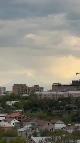 The video shows a panoramic view of a town against the backdrop of a snow-capped mountain range, under a cloudy sky.  In the foreground, there are single-family homes with slanted roofs, interspersed with trees and shrubs. In the middle ground, a few multi-story apartment buildings of brick or concrete construction dominate the view. A construction crane is visible near one of the buildings.  The background features a mountain, presumably Mt. Ararat, its peaks covered in snow and partially obscured by clouds. The sky is filled with dramatic grey clouds with the sunlight illuminating parts of the landscape and highlighting the snow. Several birds are flying around.