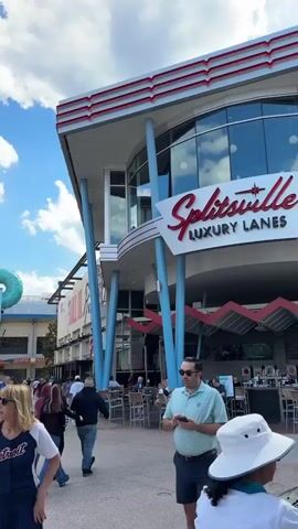 The video shows a bustling outdoor shopping or entertainment complex under a partly cloudy sky.  People of varying ages and appearances are walking along a paved pedestrian area.  In the first few seconds, a woman wearing a Detroit Tigers baseball jersey is visible. The camera pans across the scene revealing businesses, including a Splitsville Luxury Lanes and an AMC movie theater, the latter displaying banners for "The Devil Wears Prada". Other shops and restaurants line the walkway, adding to the vibrancy of the scene. People are window shopping, walking in groups, and generally enjoying the outdoor environment. The overall atmosphere is lively and commercial.