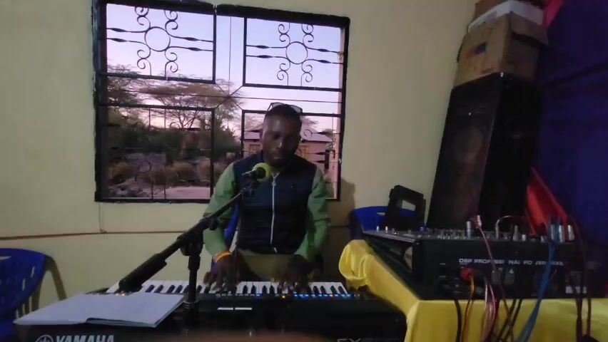 A man is sitting at a keyboard in a room. He is wearing a green shirt, a blue vest, and dark pants. A microphone is in front of him, and he is playing the keyboard.  To his right is a sound mixing board covered in a yellow cloth.  To the side of the mixing board is a large black speaker and a stack of cardboard boxes.  A window is behind him, and outside of the window is a landscape with trees and a house. There are two blue plastic chairs on the left and one behind the mixing board.  The keyboard is labeled "YAMAHA SX600".