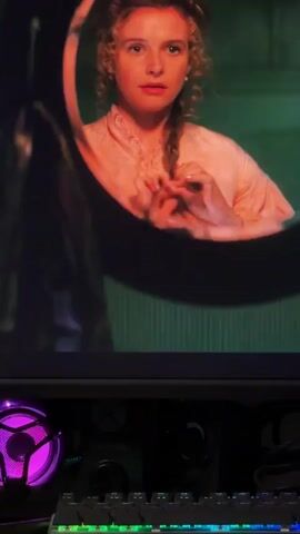 The video shows a woman with blonde hair in a pink dress braiding her hair in a mirror. A man with a beard in a vest and white shirt enters the frame and speaks to her. The background is a dark, industrial-looking space with a light fixture visible. A computer monitor displaying the scene is in the foreground.
