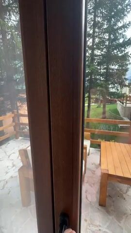 The video shows a view from inside a building looking out onto a patio area. The patio is furnished with wooden chairs and a table. There are trees and a fence visible in the background. The door appears to be opening or closing.