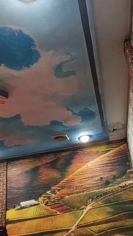 The video shows an interior shot, with a ceiling painted with a sky mural and a wall mural depicting a landscape with rolling hills and fields. The camera pans slowly from left to right, revealing parts of the murals. The ceiling features clouds in various shades of blue, pink, and white. The wall mural is vibrant, with colors of green, yellow, orange, and red representing cultivated fields and rows of trees or vines. A brick pillar is visible on the right side of the frame. A light fixture is visible on the ceiling. There are no people or discernible actions taking place in the video.