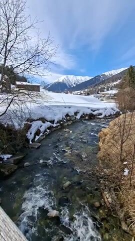 A clear, shallow river flows swiftly over rocks, its banks lined with patches of snow. Beyond the snow-covered fields, a small village nestles at the foot of snow-capped mountains under a bright blue sky with scattered clouds. Leafless trees stand on either side of the riverbank.
