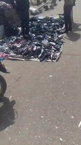 The video shows a pile of used shoes laid out on a tarp on the ground. Several people are standing around the shoes, presumably looking at them or buying them. The scene appears to be a market or roadside sale of used footwear. The camera then pans to show a street with a gate that says "WELCOME TO TOFO" above it, and motorcycles are driving through the gate.  The environment is outdoors, seemingly in a public area.