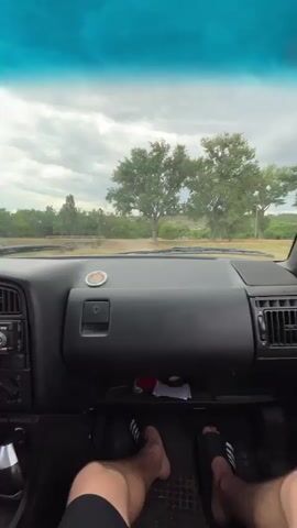 The video is a first-person perspective from inside a car. The camera is pointed forward through the windshield at a dashboard and the exterior landscape. The dashboard is dark, with a radio, vents, and a glove compartment visible. There are two feet wearing slide sandals in the foreground, suggesting the person is driving. Outside, there are trees and a cloudy sky. The car appears to be moving on a dirt road.