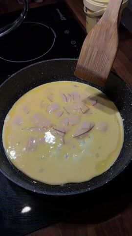 The media shows a close-up of a frying pan on a stovetop. The pan contains scrambled eggs and pieces of ham, with a wooden spatula being used to stir the mixture. The scene appears to be taking place in a kitchen, with a wooden countertop and a yellow container visible in the background. The lighting suggests it could be daytime or evening. The activity is cooking an omelet or scrambled eggs with ham. There is no visible text in the media. The location context is Słupsk, Poland.
