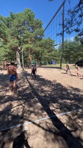 A group of people are playing volleyball on a sandy court surrounded by pine trees. The sky is clear and blue. A net is set up in the middle of the court. Two men and two women are visible playing the game. One man is in the foreground, wearing blue shorts. Another man is further back, near the net, also in shorts. One woman is in the middle of the court, wearing a black tank top and shorts, with her arms raised as if to hit the ball. Another woman is to the right, in shorts, crouching down. The ball is in the air, near the woman in the black tank top. The sun is shining, casting shadows from the trees onto the sand.