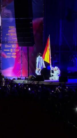 A live performance is taking place on a stage. Two individuals are visible on stage. One person, dressed in a white astronaut-like suit, is holding a Spanish flag. The other person, wearing a white outfit with a blue collar and tie, stands facing the first individual. The stage is lit with blue and pink lights, and a large speaker is mounted overhead. A crowd of spectators is visible in the foreground, many holding up their phones to record the event. The background features stage lighting and equipment.