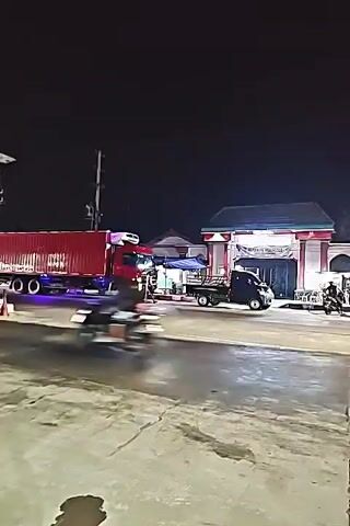 A red refrigerated truck is parked on the left side of the road. A small black flatbed truck is parked next to it. Several people are standing around the trucks. In the background, there is a building with a red and white arched entrance. There are also several motorcycles and cars parked in front of the building. The scene appears to be a roadside stop or a loading/unloading area at night. The road is wet, suggesting recent rain or a wet surface. A street light illuminates the area.