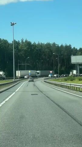 The video shows a view from a vehicle driving on a highway. The road has multiple lanes, separated by white lines. There are tire tracks visible on the asphalt, indicating recent traffic. On the left side of the road, there is a large lamppost and a metal barrier. Beyond the barrier, a dense forest of tall green trees lines the horizon.

Several vehicles are present on the highway. A large white truck with a trailer is prominently featured, appearing to be moving slowly or stopped. Other cars are also visible, some in front of and behind the truck, suggesting moderate traffic. A red and white triangular warning sign is visible on the right side of the road, indicating a hazard or a change in the road ahead. Another traffic sign with a blue circle and a white arrow pointing right is visible on the left.

The sky is a clear, bright blue with a few wispy white clouds. The lighting suggests it is daytime, likely mid-morning or afternoon, given the intensity of the sunlight. The overall impression is a typical highway scene in a wooded, possibly rural, area. No people are clearly visible within the vehicle or in other vehicles, but the presence of traffic implies human activity. The scene is peaceful and functional, with no obvious signs of distress or urgency.