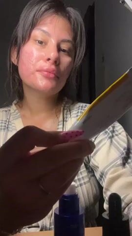 A young woman with dark hair and visible facial redness is in a dimly lit room, wearing a plaid robe. She holds a tube of skincare product with a yellow label and pink accents, which she is inspecting. In front of her are two bottles, one dark blue with a white pump top and another dark colored with a dropper. Her fingernails are painted with pink and white designs.