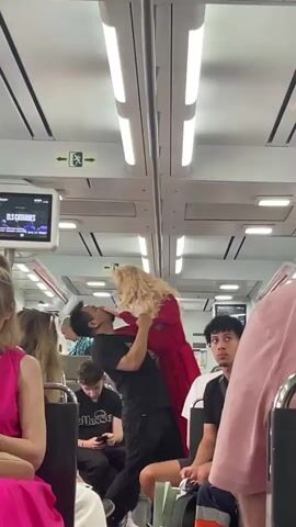 Inside a public transport vehicle, likely a train, a man in a black t-shirt and pants is holding and interacting with a large blonde doll dressed in a long red gown. He holds the doll close, and at one point, its head detaches and swings backward before he quickly reattaches it. The man continues to hold the doll upright as other passengers watch, some looking surprised or amused. The train car has overhead lighting and seats with various passengers. A screen in the background displays text including "ELS CATALANS," "QUESTA," and "DOCTOR PANTS." An exit sign with a left-pointing arrow is also visible. The scene appears to take place during the day.