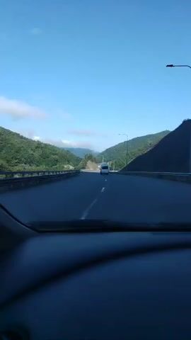 The video shows a point-of-view shot from inside a car driving on a highway. The highway is surrounded by lush green mountains under a clear blue sky with a few scattered clouds. A white van is visible driving ahead on the highway. The dashboard of the car is visible in the foreground. The camera is panning slightly to the right.