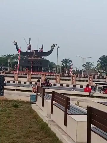 The video shows a wide shot of a roundabout with a monument in the background. There are several motorcycles and a car driving around the roundabout. A young woman is sitting on a bench in the foreground. The sky is overcast. The monument in the background appears to be a statue of two figures on a boat. There are flags lining the roundabout. The overall scene is of a public space in what appears to be a city or town.