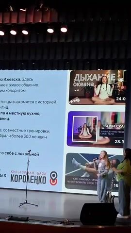 Here's a concise description of the video:

The video captures a presentation on a stage. A young woman stands at a microphone on the right side of the stage, seemingly giving a presentation. Beside her is an adult woman. A large screen behind them displays the content of the presentation, including the title "Улон" and text describing it as a female community from Izhevsk. There are also images related to the presentation shown on the screen. The stage has several microphones and speakers positioned across it. Above the stage are spotlights. The presentation content also mentions "Президентский фонд культурных инициатив" (Presidential Fund for Cultural Initiatives) and "Культурная база Короленко" (Cultural Base Korolenko).