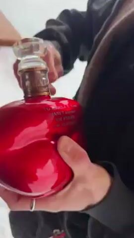 A person stands outdoors in the snow, holding a glossy, red, heart-shaped bottle. The bottle has a glass stopper. The person's hands are visible, adorned with rings. They are manipulating the stopper, possibly attempting to open the bottle. The person is wearing a dark jacket. A wooden surface is visible in the blurred background.