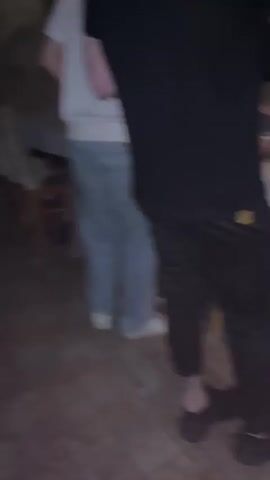 The blurry video shows several people at what appears to be an event. The camera is shaky and the focus is unclear.  A person wearing a white shirt and blue pants walks by.  Another person is wearing a white shirt and light-colored pants, and dark shoes. There is a table covered with a white tablecloth, and some bottles and other items are on the table. There are lights strung up in the background.