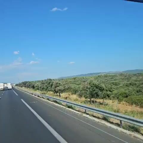 The video shows a view from inside a vehicle traveling on a highway on a sunny day. The highway has multiple lanes, and there is traffic consisting of cars and trucks. The road is flanked by greenery, with trees and bushes lining the sides. The sky is blue with a few scattered clouds. The camera pans slightly, offering a dynamic perspective of the passing scenery. The video captures the movement of vehicles on the highway and the surrounding landscape.