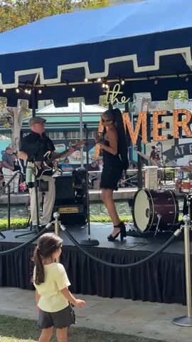 A young girl with dark hair pulled back watches a live band perform on an outdoor stage. The band is positioned under a blue and white striped awning. A female singer in a black sequined dress and high heels stands center stage, holding a microphone. To her left, a man in a cap and light-colored pants plays a guitar. To her right, a drummer in a black shirt and cap plays a drum set. Behind the band, large illuminated letters spell out "the AMERIC". The stage is enclosed by black fabric draping. In the background, a body of water is visible, with people seated on the far side. The overall setting appears to be a sunny outdoor venue.