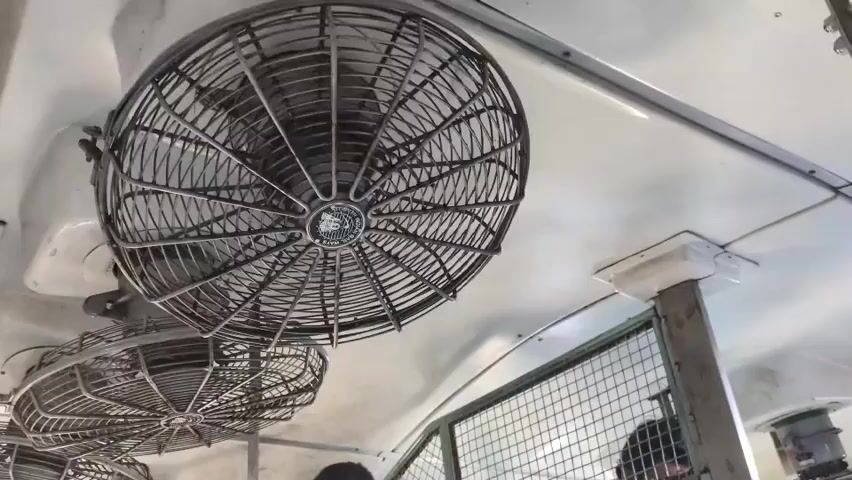 A close-up shot shows the ceiling of a vehicle with several industrial-style fans attached. The fans are dark metal with protective cages. The fan in the foreground is larger and positioned centrally, while another smaller fan is partially visible below it. The ceiling is white and there are some white supports and metal bars visible. The video seems to capture the fans in motion.