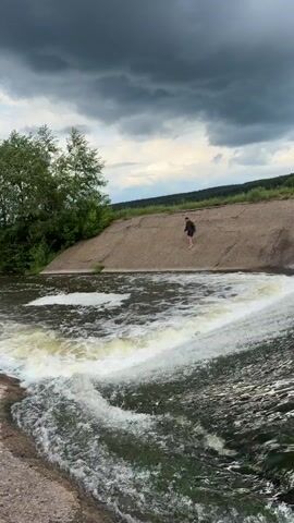 The video shows a man walking on a concrete structure next to a small waterfall. The waterfall spills into a wide, rushing river. The man is wearing dark clothing and appears to be barefoot. The sky is overcast with dark, stormy clouds. There are trees and vegetation along the riverbank. The scene is mostly natural, with the exception of the concrete structure.