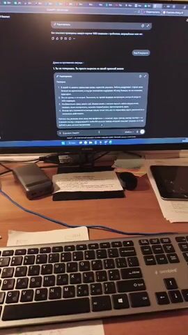 The video shows a desk with a computer monitor, keyboard, and various office supplies. On the monitor, a chat interface with the heading "ChatGPT" is visible. The chat displays a conversation in Russian, with the user asking a question about overcoming the 1000 limitations of "superhuman intelligence" and the bot providing a detailed response. The user's query is titled "The limitations of "superhuman intelligence", and the bot's response is titled "Response". The response is presented in a numbered list format. The desk also has a smartphone, a power bank, and some papers. The background shows a tiled wall with several sticky notes attached. The lighting suggests an indoor environment, likely an office or study.