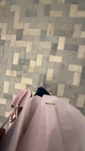The video shows a first-person perspective of someone walking on a patterned tiled floor. The person is wearing a light-colored skirt or dress, dark shoes with a colorful pattern, and carrying a light-pink purse and a magazine. The camera is angled downwards, focusing on the person's legs and feet as they walk. The environment appears to be outdoors or in a covered walkway, as indicated by the lighting and the tiled ground. The person is walking at a normal pace.