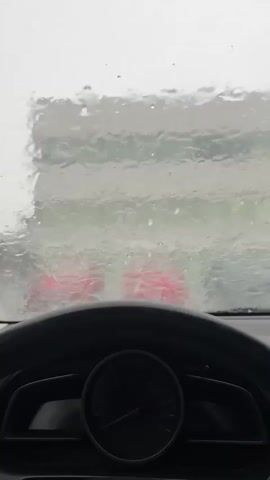 From the perspective inside a car, the windshield is heavily obscured by rain. Two red lights are vaguely visible through the rain-streaked glass, suggesting the presence of another vehicle. The car's steering wheel and dashboard, including the speedometer, are visible in the foreground. The environment appears to be overcast and rainy.